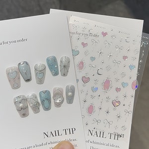 May include: A set of nail art stickers with a variety of designs, including hearts, stars, bows, and moons. The stickers are silver and have a holographic finish. The text "NAIL TIP" is printed on the sheet.