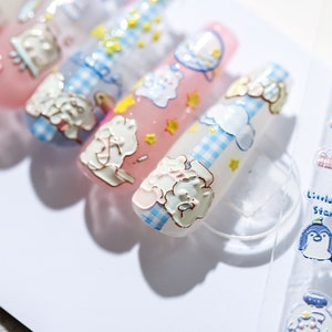 May include: Close-up of several long, decorated fingernails. The nails feature cartoon bear and cloud designs in white, blue, and pink. Some nails have a gingham pattern. Yellow star accents are also present. The overall theme is whimsical and cute.