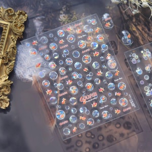 May include: Clear nail art stickers featuring goldfish swimming in bubbles. The stickers have the word "Goldfish" and the phrase "Carpe diem" printed on them. The stickers are transparent with orange and yellow fish.