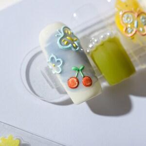 May include: A white fake nail with a blue butterfly, a white flower, and two red cherries painted on it.