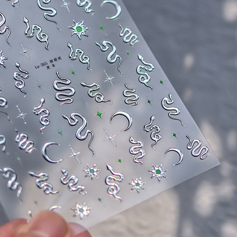 May include: A sheet of silver nail stickers with a variety of designs, including snakes, moons, stars, and suns. The stickers are on a clear background and have a glossy finish.
