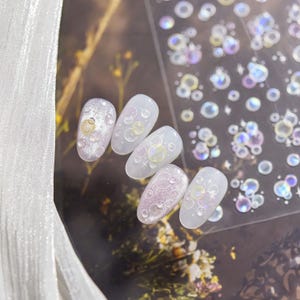 May include: A set of artificial nails with a bubble design. The nails are oval-shaped and feature a variety of iridescent and clear bubble accents. Some nails have a glittery base, while others are clear. A sheet of bubble stickers is also visible.