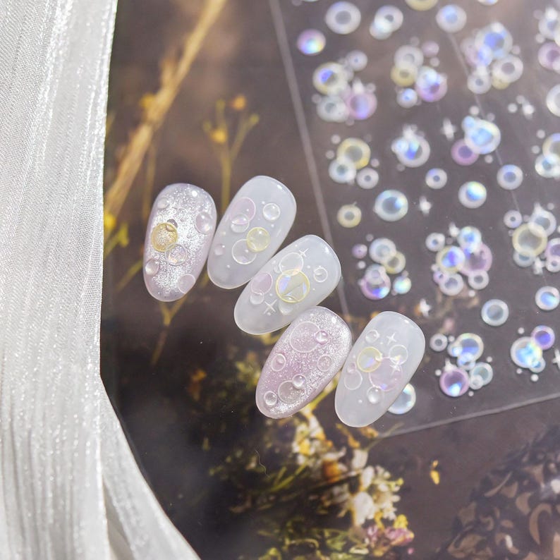 May include: A set of artificial nails with a bubble design. The nails are oval-shaped and feature a variety of iridescent and clear bubble accents. Some nails have a glittery base, while others are clear. A sheet of bubble stickers is also visible.