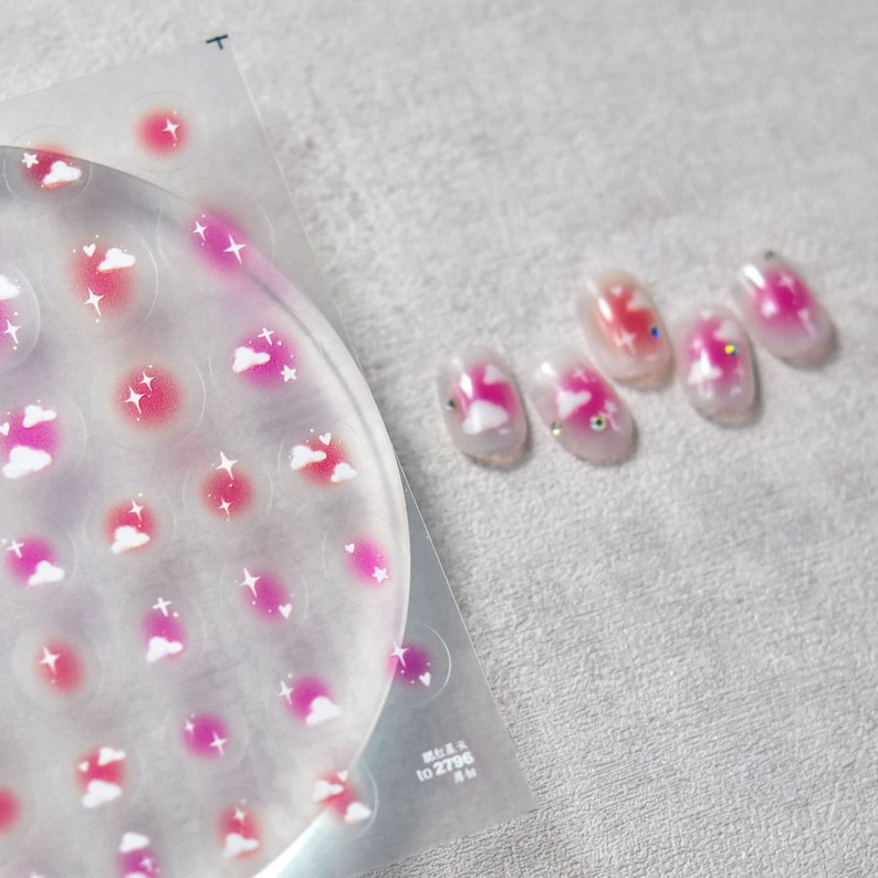 May include: A sheet of nail stickers with a pink and white cloud and star design. The sheet is clear and has a circle of stickers in the center. There are five nails with the same design on a white surface.