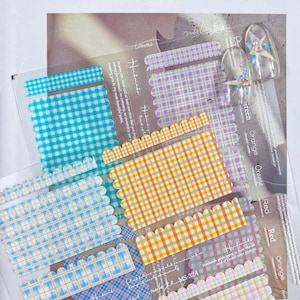 Puede incluir: Una colección de pegatinas para uñas con varios patrones de cuadros, incluyendo azul, morado y naranja. Las pegatinas tienen un borde festoneado y se muestran en una lámina transparente. También se ve una uña decorada con una flor y un lazo.