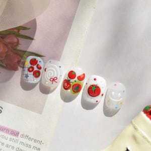 May include: A set of five artificial nails with various designs. The nails feature tomato and smiley face designs, with red, green, and white colors. Some nails have a grid pattern, swirls, and stars.
