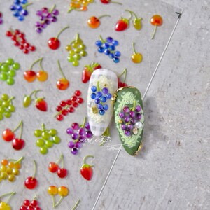 Pu&ograve; includere: Sticker per nail art con diversi motivi di frutta, tra cui ciliegie, fragole, uva e uva verde. Gli adesivi sono disposti su uno sfondo bianco con un motivo a scacchi.