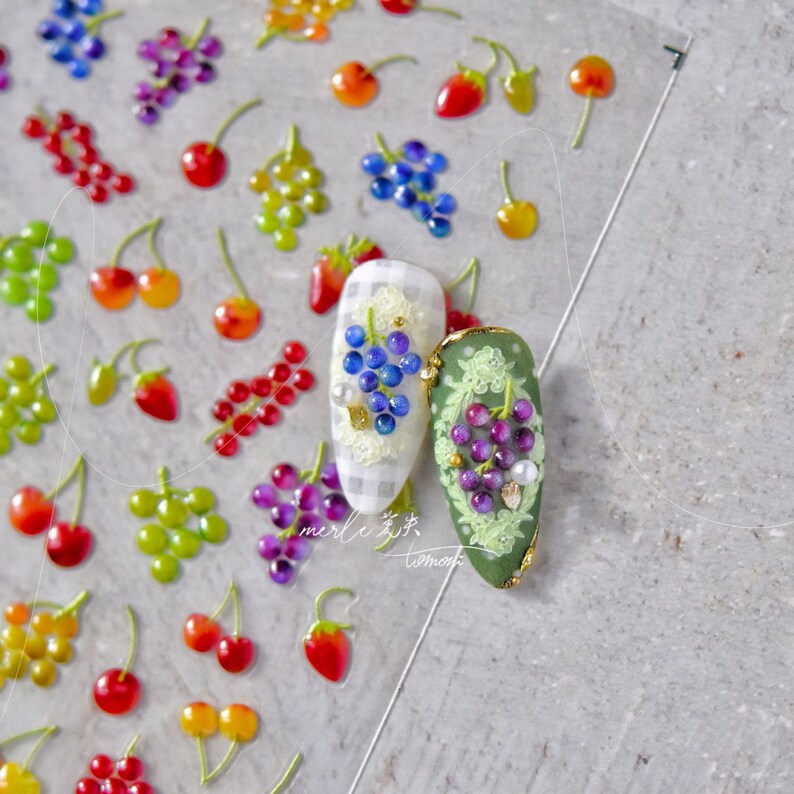Pu&ograve; includere: Sticker per nail art con diversi motivi di frutta, tra cui ciliegie, fragole, uva e uva verde. Gli adesivi sono disposti su uno sfondo bianco con un motivo a scacchi.