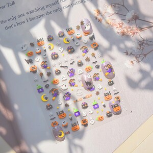 Peut inclure: Une feuille d'autocollants pour ongles sur le th&egrave;me d'Halloween avec une vari&eacute;t&eacute; de motifs, notamment des citrouilles, des chats, des fant&ocirc;mes, des chauves-souris et des &eacute;toiles. Les autocollants sont sur un fond violet et ont une finition brillante.