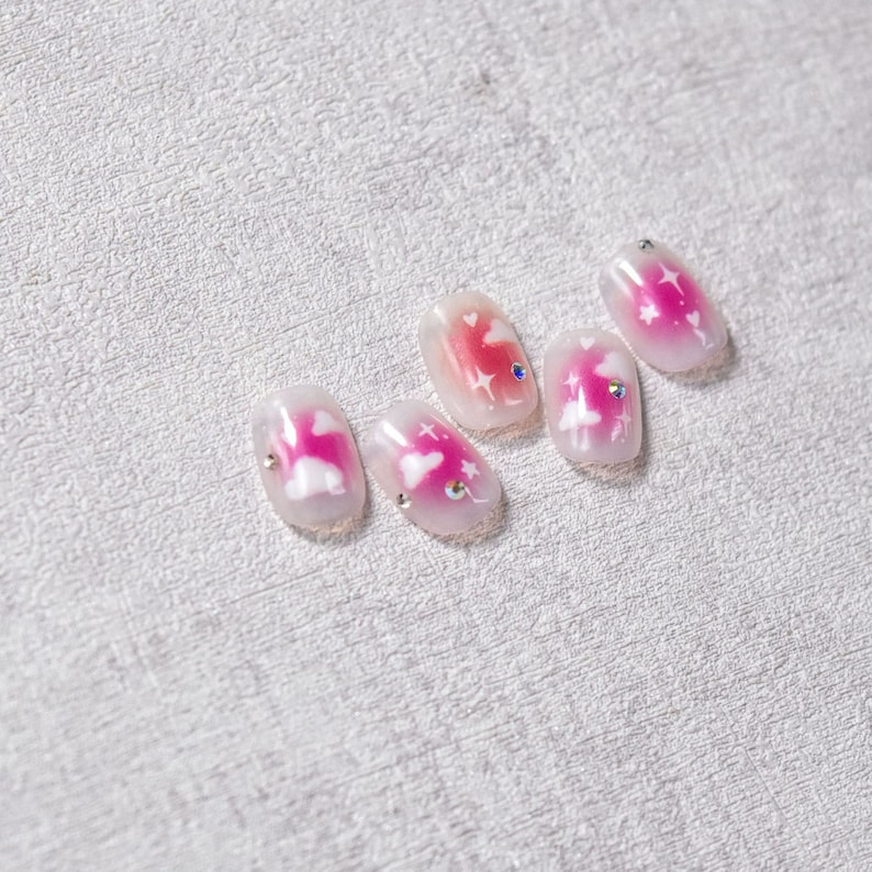 May include: Five pink and white fake nails with a cloud and star design. Each nail has a small clear rhinestone.