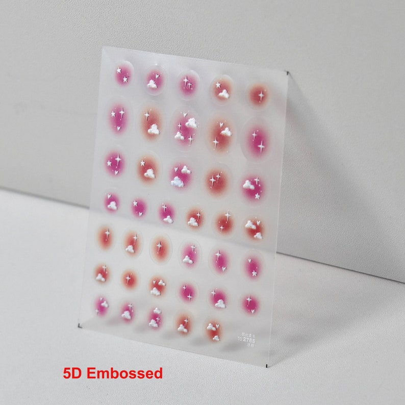 May include: A sheet of 5D embossed nail stickers with a design of pink and orange clouds with white stars. The stickers are on a clear sheet.