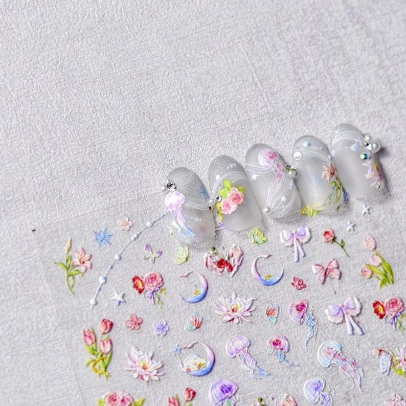May include: A sheet of nail art stickers featuring a variety of designs, including flowers, moons, butterflies, and jellyfish. The stickers are on a clear background and are ready to be applied to nails.