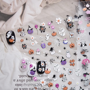 May include: Halloween-themed nail art stickers featuring pumpkins, ghosts, spiders, and witch hats. The stickers are in orange, purple, white, and black. The image also shows several decorated fingernails with similar designs.