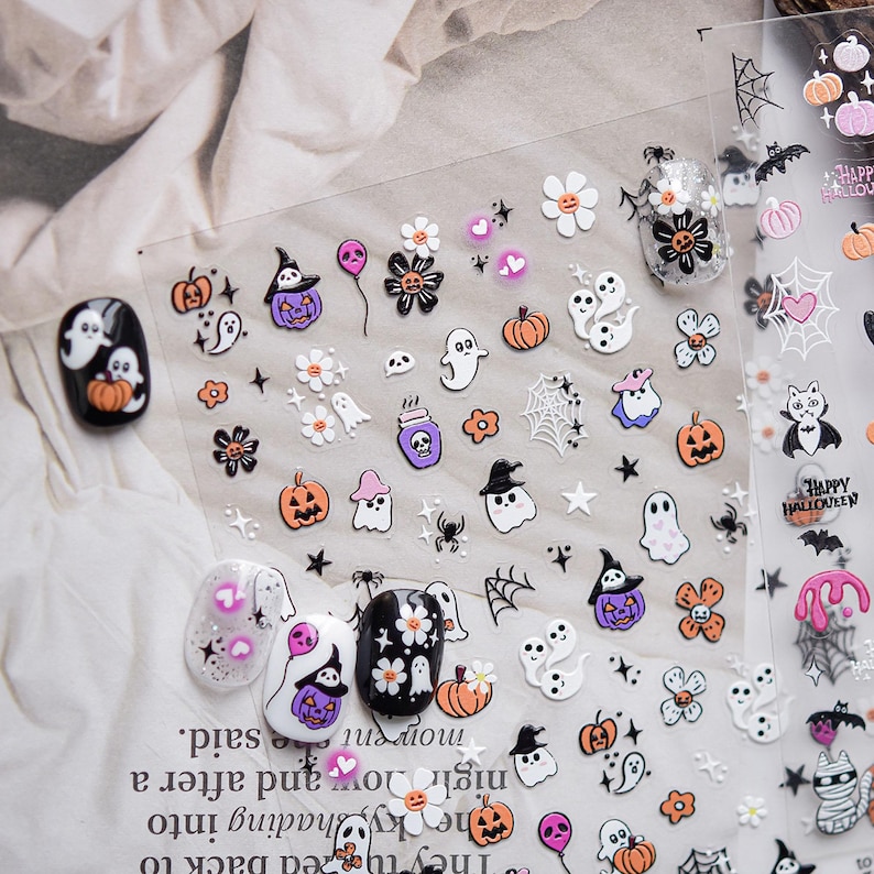 May include: Halloween-themed nail art stickers featuring pumpkins, ghosts, spiders, and witch hats. The stickers are in orange, purple, white, and black. The image also shows several decorated fingernails with similar designs.