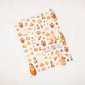 Puede incluir: Una hoja de pegatinas para u&ntilde;as con tem&aacute;tica oto&ntilde;al con ardillas, calabazas, hojas, bellotas y rosquillas. Las pegatinas tienen el texto "Good Day", "Falling Leaves", "Hello Autumn", "Golden Pumpkin" y "Let's go for a walk".