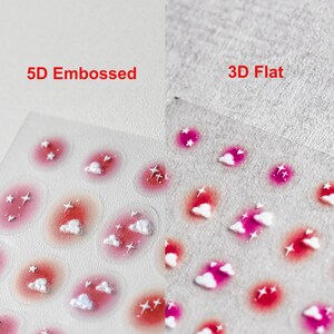 May include: Two sheets of nail stickers with a pink and white cloud design. The left sheet shows the stickers in a 5D embossed style, while the right sheet shows the stickers in a 3D flat style. The text "5D Embossed" and "3D Flat" is printed above each sheet.