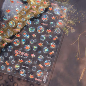 May include: Clear sticker sheet featuring orange goldfish and bubble designs. The sheet includes the word "GOLDFISH" and the phrase "Carpe diem." The stickers have a holographic effect, creating a shimmering appearance.