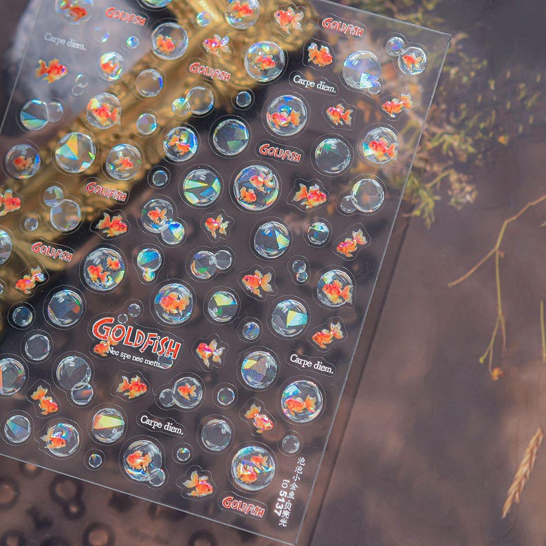 May include: Clear sticker sheet featuring orange goldfish and bubble designs. The sheet includes the word "GOLDFISH" and the phrase "Carpe diem." The stickers have a holographic effect, creating a shimmering appearance.