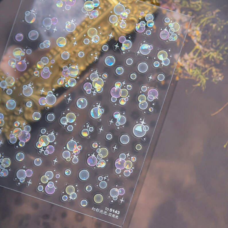 May include: Clear sheet of stickers featuring iridescent bubble and star designs. The stickers have a holographic effect, with colors shifting from blue to yellow and purple. The sheet also includes the text "TO 5143" and Chinese characters.