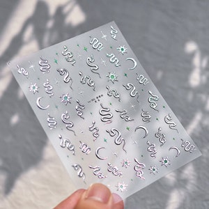 May include: Silver and green snake, moon, and star nail stickers. The stickers are on a clear sheet.