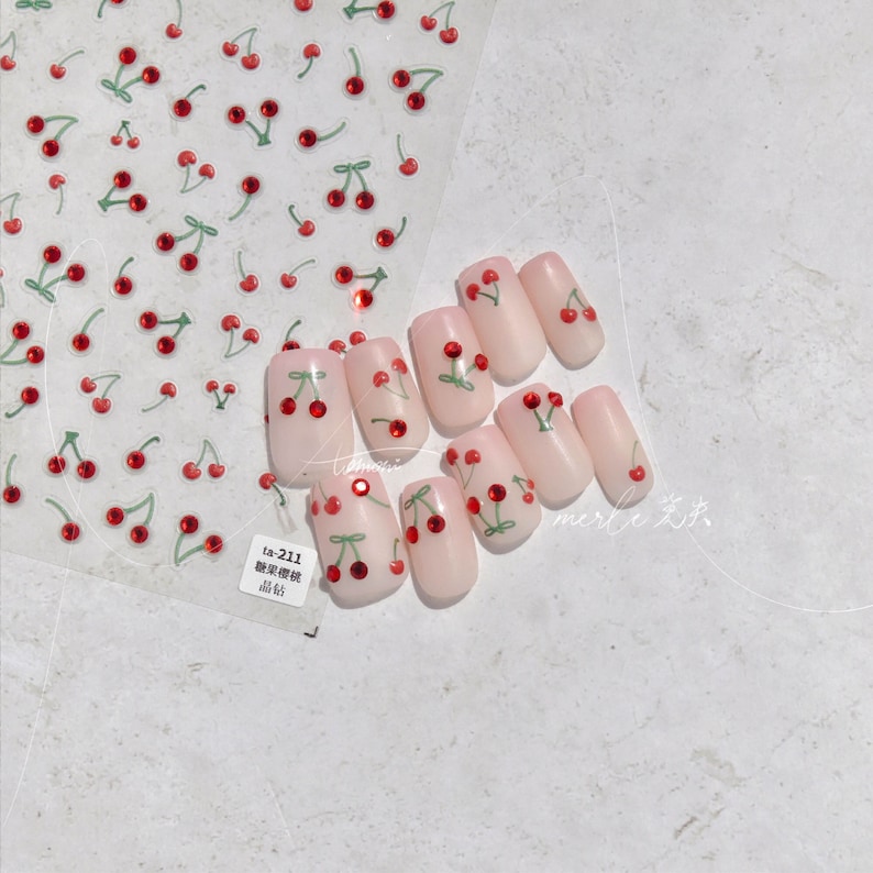 May include: A set of false nails with a cherry design. Each nail is painted a light pink color and has a small cherry with a green stem and red fruit. The cherries have a single rhinestone in the center. The nails are arranged in a row on a white background. The image also includes a sheet of nail stickers with a cherry design.
