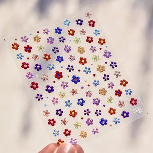 May include: A sheet of colorful flower-shaped stickers with a rainbow of colors including red, orange, yellow, green, blue, purple, and pink. The stickers are on a clear background.