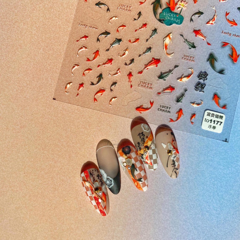 May include: Nail art stickers with koi carp designs in red, orange, and green, and the words "Lucky Charm". Five decorated fingernails are also visible, with designs including checkered patterns and floral accents.