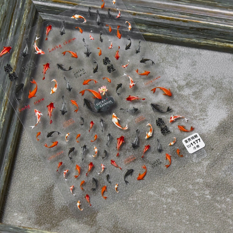 May include: Clear sheet of nail art stickers featuring koi carp in shades of red, orange, black, and white. The stickers include the text "Lucky Charm" and other Chinese characters. The sheet has a small white sticker with Chinese text.