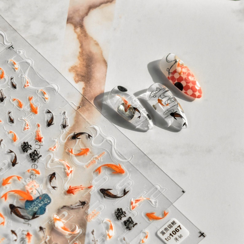 May include: Clear nail art stickers with orange, black, and white koi fish designs. The stickers are on a clear sheet with embossed wave patterns. Three manicured nails showcase koi fish designs, including a red and white checkered pattern.