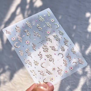 May include: A sheet of gold and pink nail stickers with a repeating pattern of snakes, moons, stars, and suns. The sheet is labeled "tu-2055"