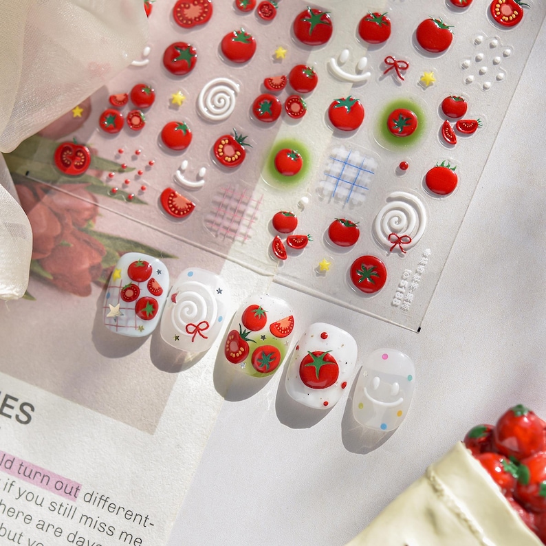May include: Clear sheet of nail art stickers featuring red tomatoes, stars, bows, and smiley faces. Several nails are decorated with tomato designs. The nail art stickers are on a white background.