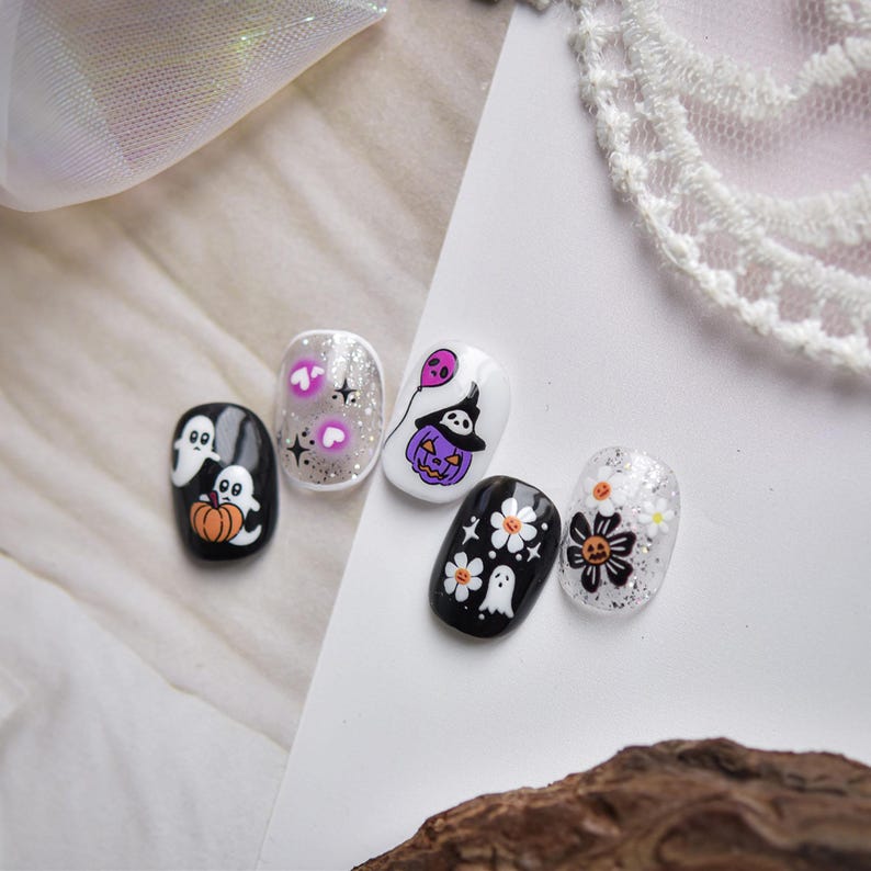 May include: A set of five Halloween-themed nail art designs. The designs include ghosts, pumpkins, flowers, and a witch's hat on a pumpkin. The nails are painted in black, white, and silver with glitter accents.