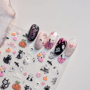 May include: Halloween-themed nail art stickers featuring pumpkins, ghosts, cats, and spiderwebs. The nail designs include the words "Happy Halloween" and various spooky illustrations in black, pink, and white. The nails are painted in black, pink, and clear colors.