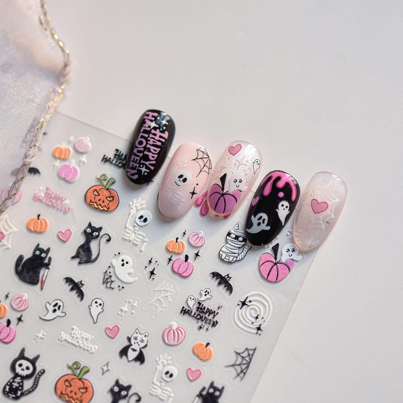 May include: Halloween-themed nail art stickers featuring pumpkins, ghosts, cats, and spiderwebs. The nail designs include the words "Happy Halloween" and various spooky illustrations in black, pink, and white. The nails are painted in black, pink, and clear colors.