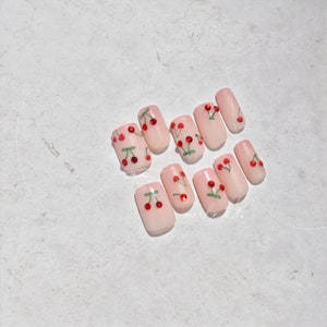 May include: Set of ten pink press-on nails with a cherry design. Each nail has a red cherry with a green stem and leaf. The cherries have a small rhinestone in the center.