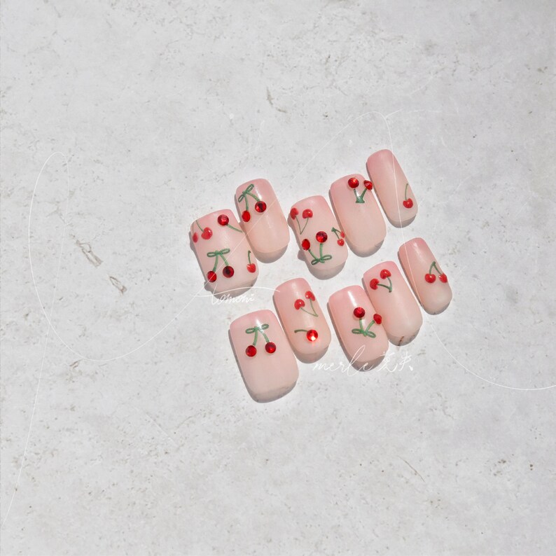 May include: Set of ten pink press-on nails with a cherry design. Each nail has a red cherry with a green stem and leaf. The cherries have a small rhinestone in the center.