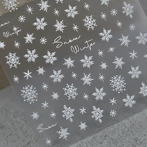 Op de afbeelding: Een vel met witte sneeuwvlok-nagel stickers met de tekst "Snow Winter" erop gedrukt.