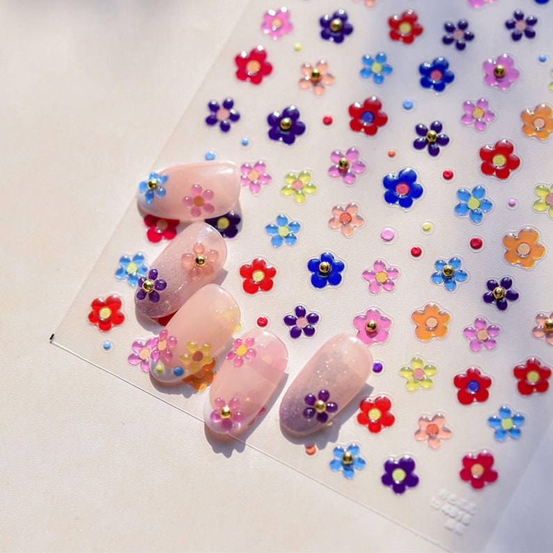 May include: A sheet of colorful flower nail stickers with a gold center. The stickers are arranged in rows on a white background. Five fingernails are shown with the stickers applied.