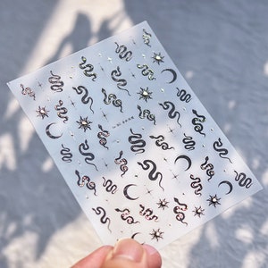 May include: A sheet of clear sticker paper with a variety of gold and black snake, crescent moon, and sun designs. The stickers are perfect for adding a touch of whimsy to journals, planners, and other craft projects.