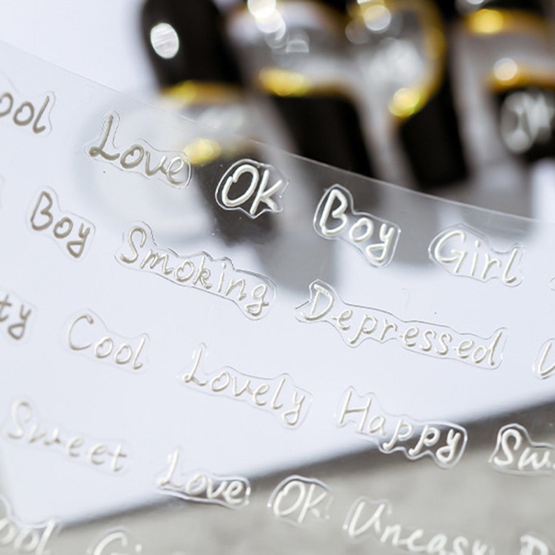 May include: A clear sheet of transparent nail art stickers with white lettering. The words on the stickers include "Love", "OK", "Boy", "Girl", "Smoking", "Depressed", "Cool", "Lovely", "Happy", "Sweet", and "Uncast".