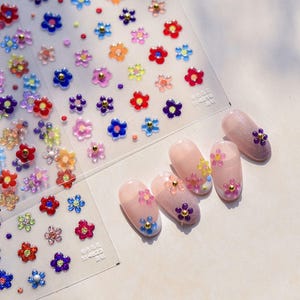 May include: A sheet of colorful flower nail stickers with a variety of colors and sizes. The stickers are shown on five fingernails with a clear base coat.
