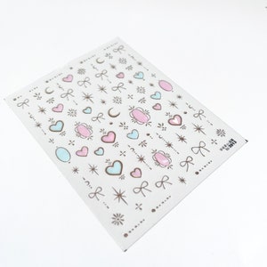 May include: A sheet of nail art stickers with a variety of designs, including hearts, stars, moons, bows, and swirls. The stickers are in shades of pink, blue, and gold.