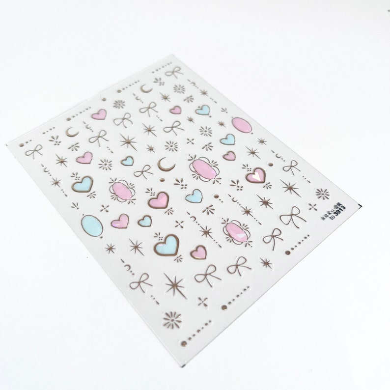 May include: A sheet of nail art stickers with a variety of designs, including hearts, stars, moons, bows, and swirls. The stickers are in shades of pink, blue, and gold.