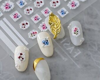 May include: Four white oval-shaped nail tips with floral designs. The nail tips have a white background with a delicate floral pattern in red, blue, and white. The nail tips are decorated with small white pearls. A gold leaf design is also visible in the image.