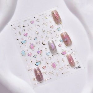 May include: A sheet of gold nail art stickers with a variety of designs, including hearts, stars, bows, and moons. The stickers are on a clear background and are ready to be applied to nails.