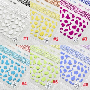 Op de afbeelding: Zes vellen met nagelstickers met een koe-print. Elk vel heeft een andere kleur: wit, geel, roze, blauw, groen en paars. De vellen zijn genummerd van 1 tot 6 en hebben de tekst "Cute Cow" erop gedrukt.
