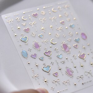 May include: A sheet of clear nail stickers with gold, pink, and blue designs. The designs include hearts, stars, moons, bows, and gemstones.