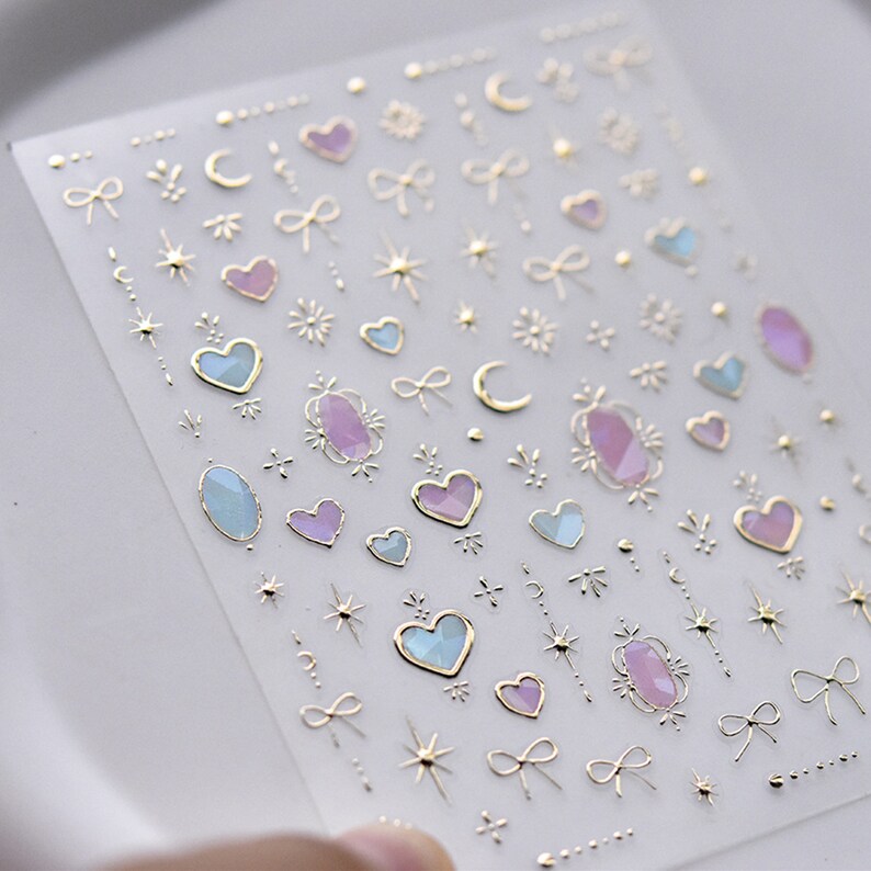 May include: A sheet of clear nail stickers with gold, pink, and blue designs. The designs include hearts, stars, moons, bows, and gemstones.