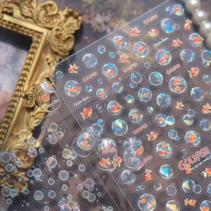 May include: Clear nail art stickers featuring goldfish in bubbles, with the words "Goldfish" and "Carpe diem." The stickers have a holographic effect, with orange and white goldfish. The stickers are on a clear sheet.