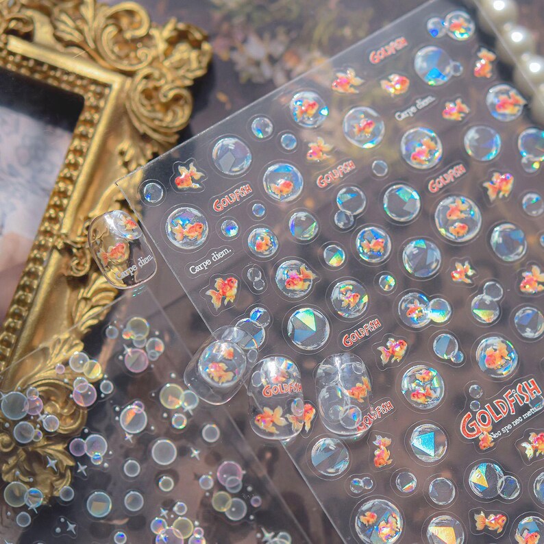 May include: Clear nail art stickers featuring goldfish in bubbles, with the words "Goldfish" and "Carpe diem." The stickers have a holographic effect, with orange and white goldfish. The stickers are on a clear sheet.